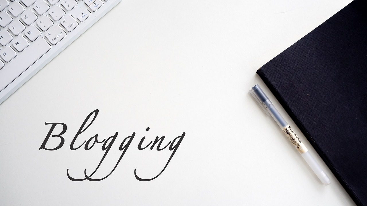 하얀 책상 위에 블로그 글쓰기 준비를 연상시키는 펜과 노트, 키보드가 놓여 있고 'Blogging'이라는 단어가 인쇄되어 있는 이미지.