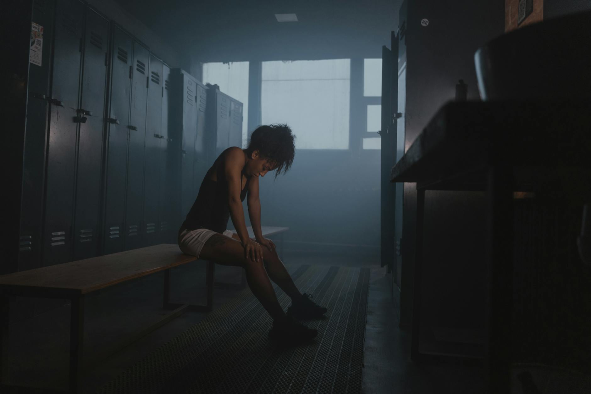 어두운 라커룸에서 지친 운동선수가 벤치에 앉아 숨 고르며 생각에 잠긴다